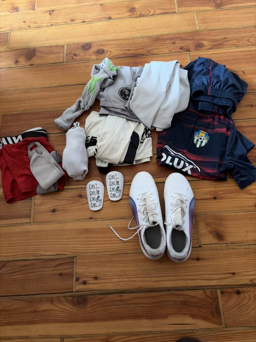 Soccer/football gear laid out on a wooden floor including white cleats with purple accents, red and white gloves, white socks, shin guards, and dark jerseys.