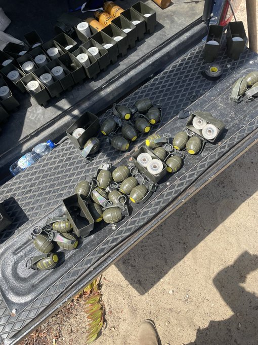An outdoor scene showing the bed of a truck loaded with numerous green military-style hand grenades and several boxes of smoke grenade canisters.