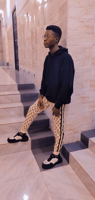 A young man wearing glasses, a black hoodie, patterned beige pants, and chunky sneakers standing on marble stairs in a building lobby.