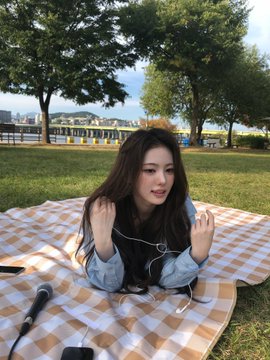 A young woman lying on a yellow and white checkered picnic blanket in a grassy park with trees and a lake in the background, holding sunglasses and wearing a light blue denim shirt.