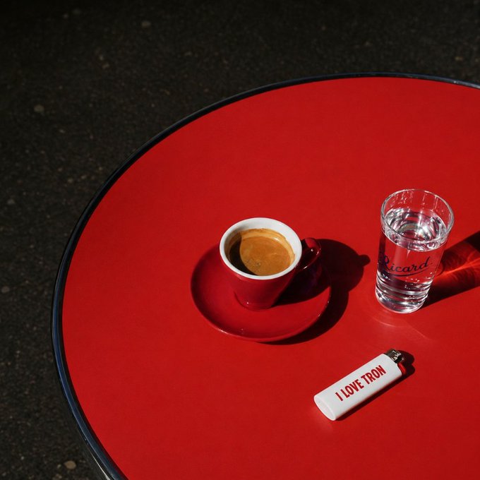 A red espresso cup and a glass of water branded "Ricard" sit on a vibrant red round table alongside a white lighter inscribed with "I LOVE TRON," perfectly matching the post's GM greeting and #TRON reference with a stylish espresso break.