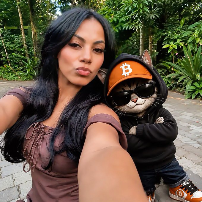 A woman takes a selfie outdoors next to an anthropomorphic cat wearing sunglasses, a black hoodie, and an orange hat with the Bitcoin symbol.