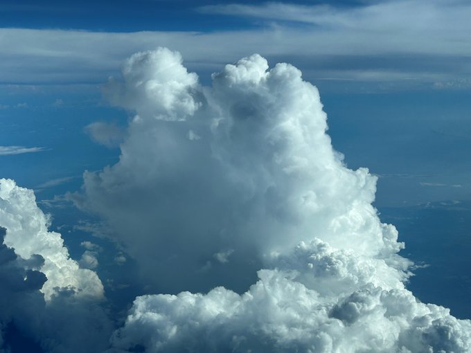 A view of large, billowing white cumulus clouds towering against a blue sky, taken from an aerial or elevated perspective.