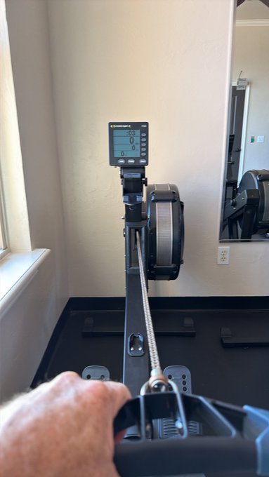 A person's hand using the handle of a Concept2 rowing machine in an indoor gym.