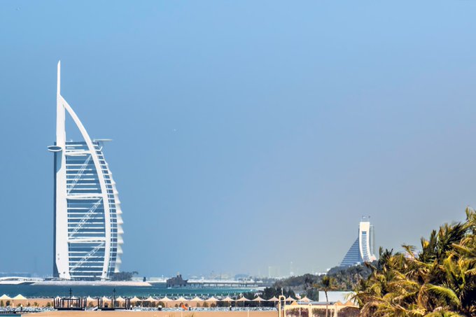 A daytime photograph of a beach scene in Dubai, prominently featuring the Burj Al Arab hotel in the shape of a sail on the left, with a building resembling the Sheraton Grand visible in the distance on the right, under a clear blue sky with palm fronds in the foreground.