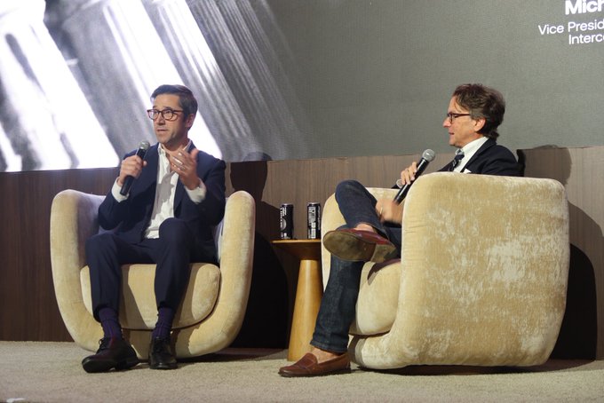 Two men sit on cream-colored chairs on a stage during an interview, both holding microphones, with 'Microsoft Vice President Interop...' visible on the screen behind them.