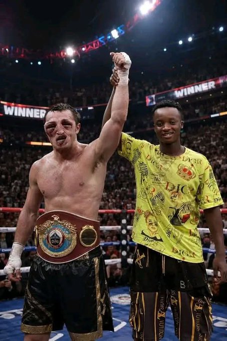 A photograph inside a boxing arena showing a victorious male boxer with his arm raised, wearing a championship belt and black and gold shorts, accompanied by a young boy in a yellow patterned shirt, with "WINNER" visible on the screens behind them.