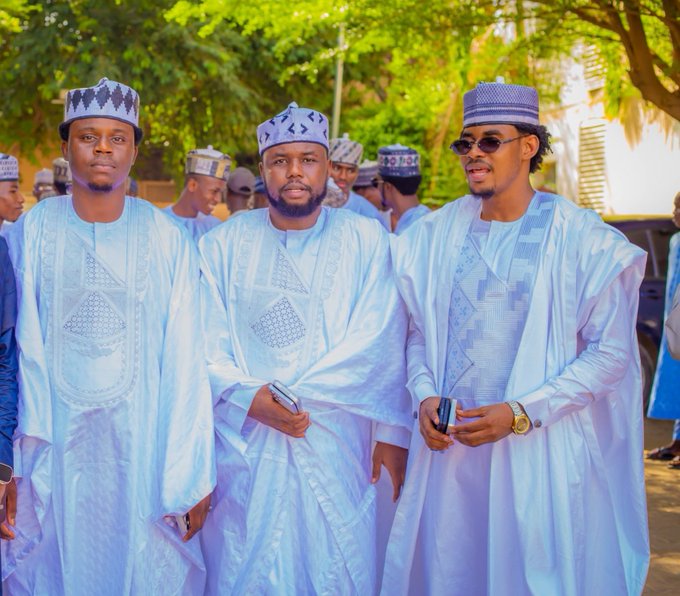 Three men in matching white traditional attire with embroidered caps stand together outdoors, posing for a photo as they appear to celebrate the completion of their work, with one man wearing stylish sunglasses and a gold watch. The image complements the post's message of gratitude to Allah for finishing their tasks and reflects the festive, communal spirit of requesting fans to tag $SUNUSI.