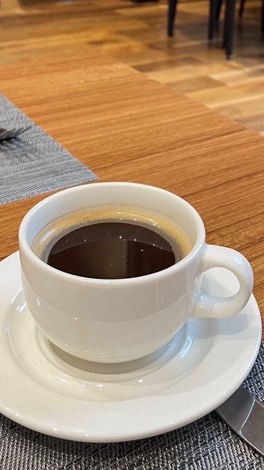 A white ceramic cup filled with black coffee sits on a saucer atop a grey placemat on a wooden table.