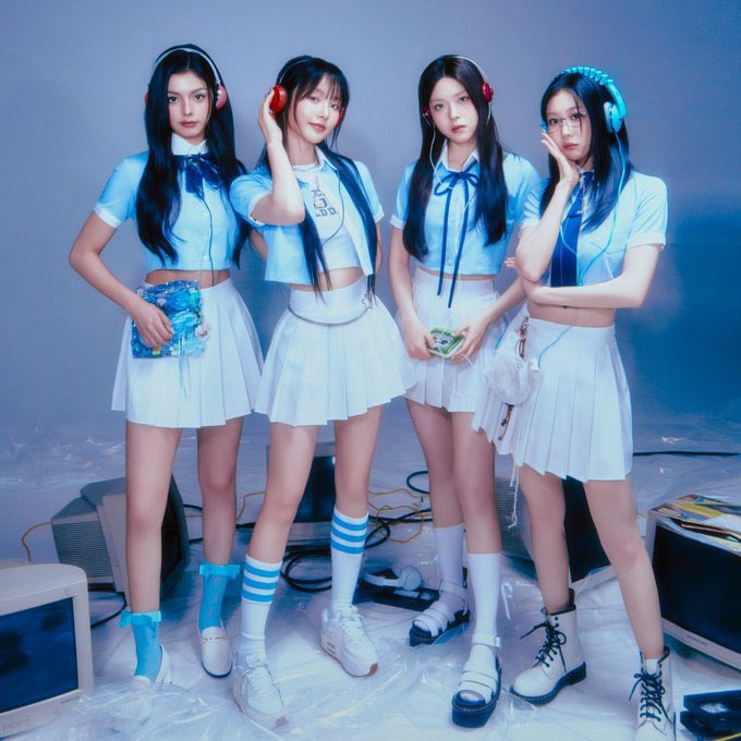 A promotional photo of four young women in matching light blue crop tops, white pleated skirts, and headphones, posing in a schoolgirl-inspired concept.