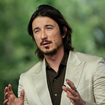 A man with shoulder-length dark hair and a goatee, wearing a cream-colored blazer over a dark shirt, gestures expressively with his hands. The image accompanies news about Robinhood ($HOOD) tokenized stocks reaching $160 million in total value.