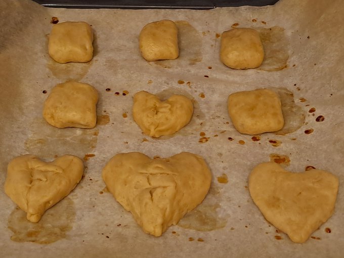 A baking tray lined with parchment paper holds nine pieces of raw dough, some shaped into squares and others into rough hearts, ready to be baked.