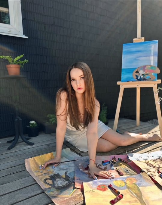A young woman in a white dress sits on a wooden deck surrounded by paintings, paint tubes, and brushes, with an easel holding a colorful seascape painting behind her.