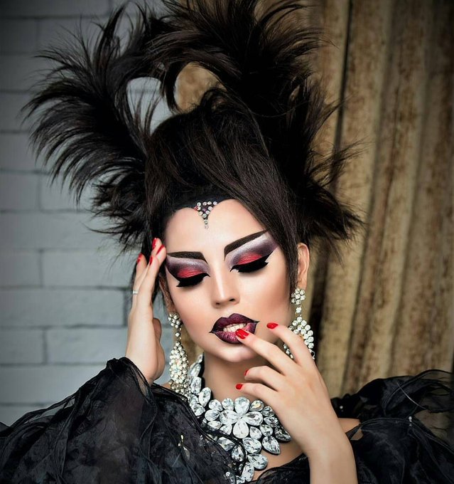 A dramatic portrait features a person with voluminous spiked black hair, bold dark eye makeup, dark lipstick, and large crystal jewelry, posed against a brick wall background.