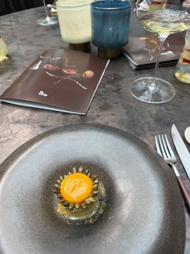 An elegant plated dish featuring a bright orange egg yolk in the center of a dark sauce with sunflower seeds, set on a gray plate next to wine glasses and a menu.