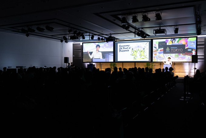A dimly lit conference auditorium where an audience faces a stage with large projection monitors; the slides feature a speaker and the text 'Economy: The Proof of Presence' alongside a logo for 'ETHGLOBAL TOKYO'.