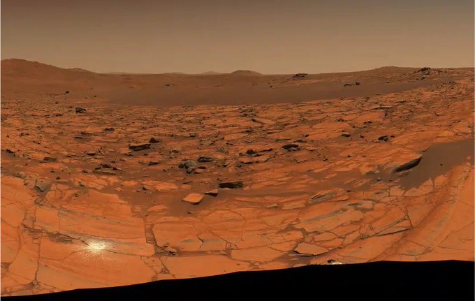 A panoramic view of the rocky, reddish-brown Martian landscape with layered sedimentary rock formations under a hazy sky.