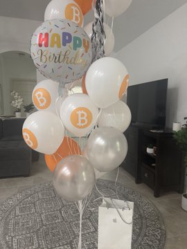 A birthday balloon arrangement featuring a "Happy Birthday" balloon and white balloons printed with orange Bitcoin "₿" symbols, tied together in a living room setting. The crypto-themed decorations match the post celebrating a BTC-themed birthday celebration.