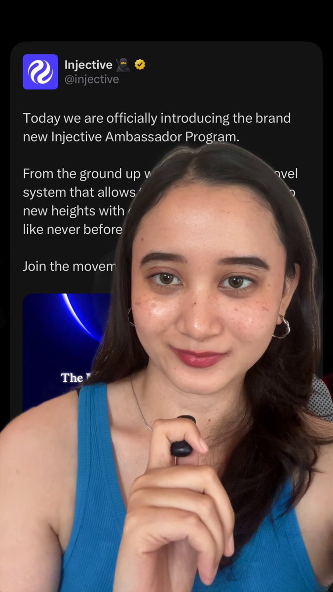 The image shows a woman wearing a blue tank top holding a black object with her right hand while looking at the camera. The background of the image is white, making the woman stand out against it. There are no other objects or people visible in the image.