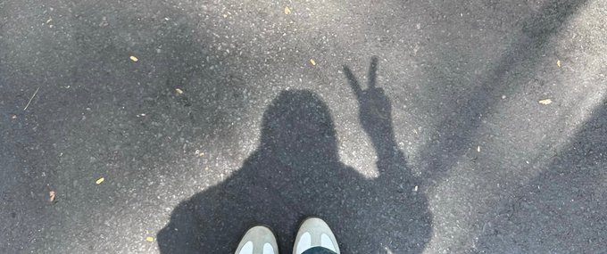 The image shows a person's shadow cast on the ground, with their feet visible above them. The shadow appears to be of a person wearing white shoes, holding up two fingers as if making peace or showing support for something. The background features a gray pavement, and there is text in the image that reads "gm good morning let's make Tuesday count @NomismaNetwork @useTria". This suggests that the person with their shadow might be promoting positivity or supporting an idea on this particular day.