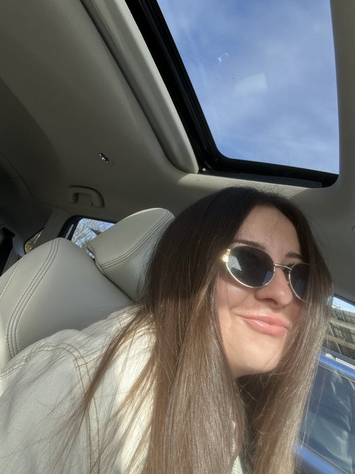 A selfie of a smiling woman with long brown hair wearing sunglasses, sitting inside a car with the sunroof open to a blue sky.