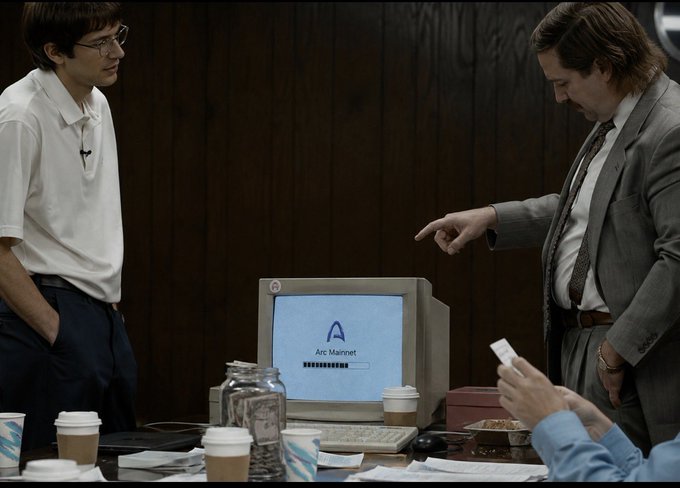 A scene from a film/TV show featuring men in business attire gathered around a vintage CRT computer monitor displaying a login screen for "Arc Manneft," with coffee cups on the table, in a wood-paneled room.