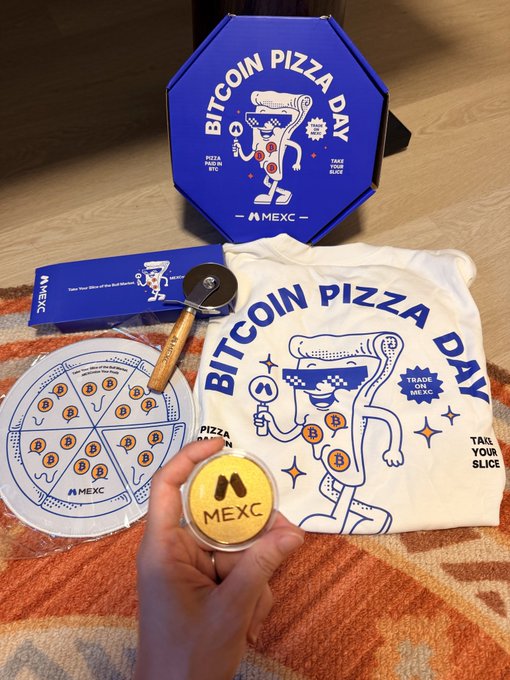 The image features a hand holding up a pizza box, which has a pizza inside of it. The box also contains a pizza cutter and a pizza slice. In addition to these items, there are two pizzas placed next to each other on a table or floor. One pizza is labeled "Bitcoin Pizza Day" with a cartoon character design, while the other pizza is not labeled at all. A person's hand can be seen holding one of the pizza boxes in the image.