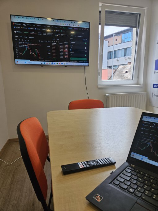 A small office or meeting room with a TV mounted on the wall displaying a cryptocurrency trading chart, a laptop on a wooden table also showing trading charts, two orange chairs, a window, and a remote control on the table.
