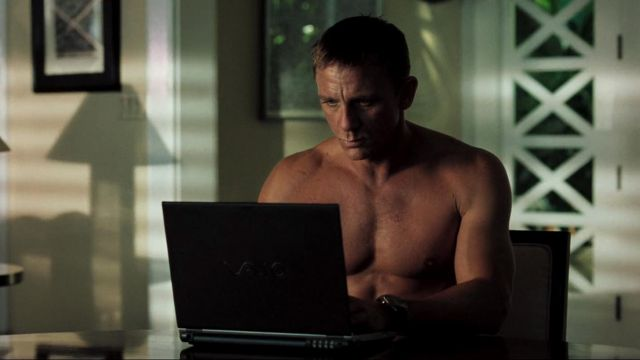 A shirtless man sits at a table intently focused on a Sony VAIO laptop. Sunlight filters through a patterned window in the background of the dimly lit room.