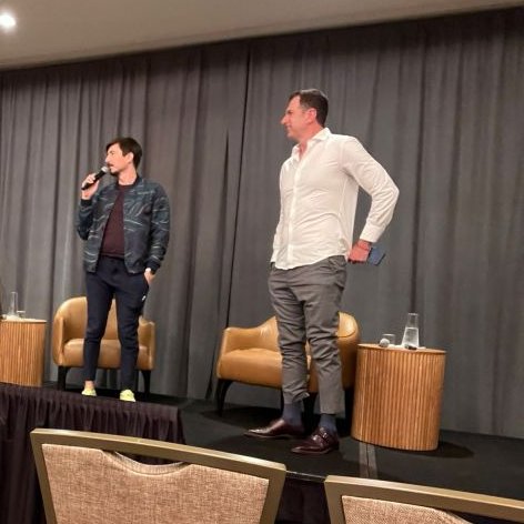 Two men speak on stage at what appears to be a conference or panel discussion, one holding a microphone while standing near empty chairs and small tables with water glasses. The setting reflects the post's theme about crypto founders discussing investment opportunities, with the speakers likely sharing insights on token economics and retail investor advantages.
