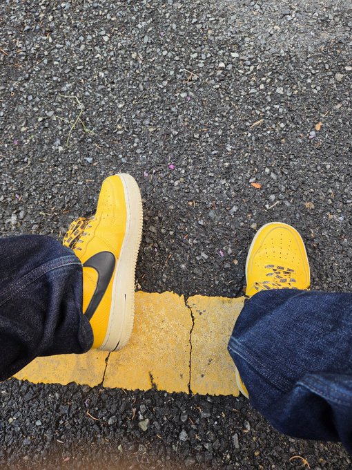A first-person perspective looking down at a pair of yellow Nike sneakers with distressed detailing, worn with blue jeans, standing on asphalt next to a cracked yellow painted line.