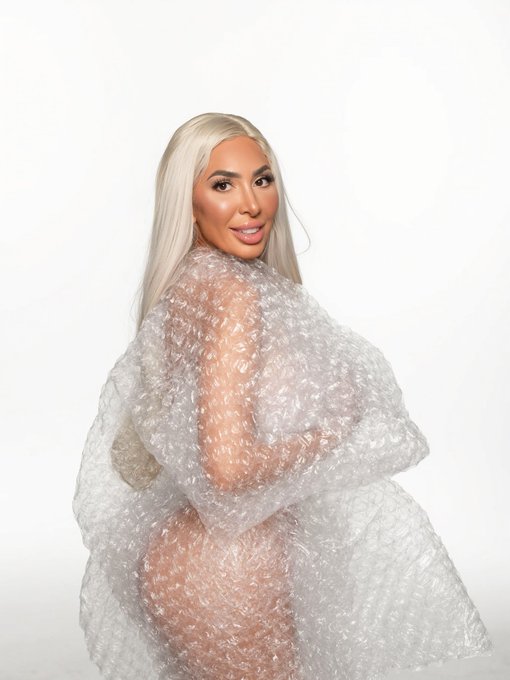 A blonde woman poses against a white background wrapped in clear bubble wrap, wearing heavy makeup.