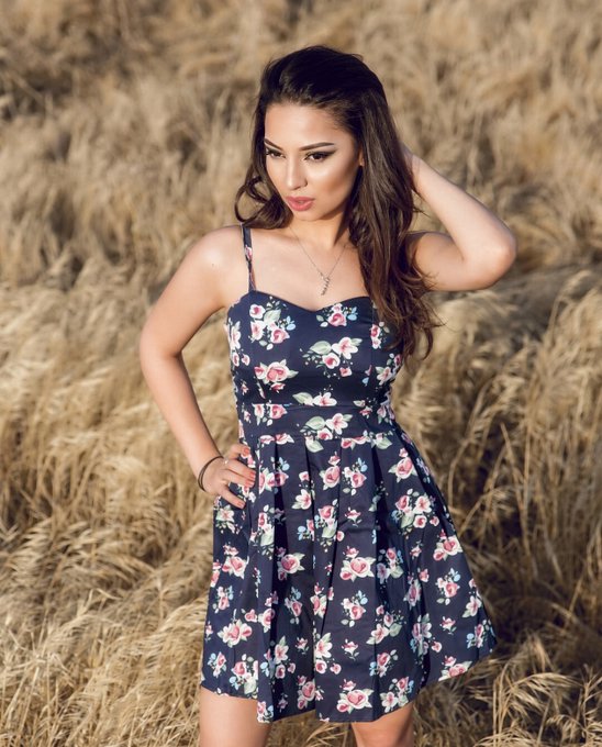 A woman with long brown hair poses in a navy blue floral dress against a backdrop of tall, dry golden grass.