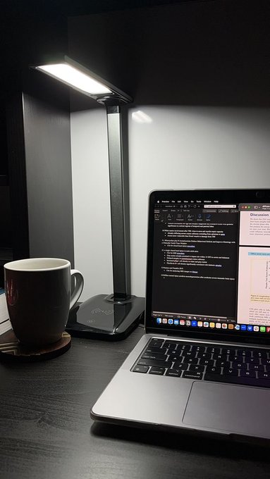 A modern desk setup featuring a sleek LED lamp illuminating a laptop screen displaying a document, accompanied by a dark coffee mug on a wooden coaster.