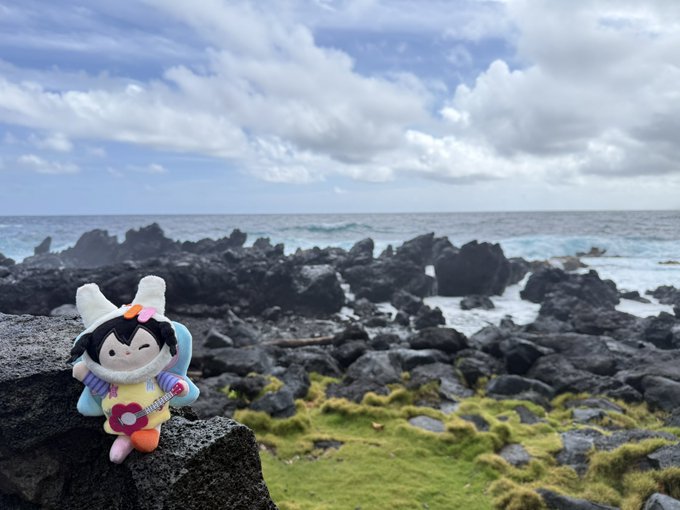 A small plush doll character sits perched on a dark volcanic rock along a rugged, algae-covered coastline with crashing waves under a partly cloudy sky, capturing a travel moment on Hawaii's Big Island. The scenic ocean backdrop complements the post's hashtag #ANYaroundtheworld, showcasing the toy's journey to a beautiful seaside destination.
