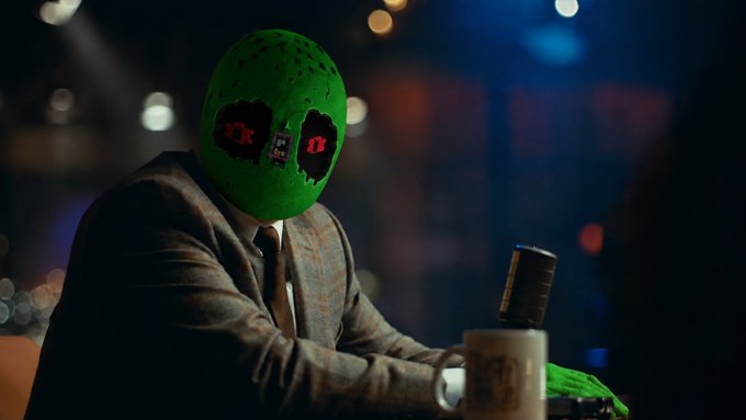 A figure in a suit and tie sits at a bar, wearing a green hockey mask with glowing red eyes, with a coffee mug visible on the counter.