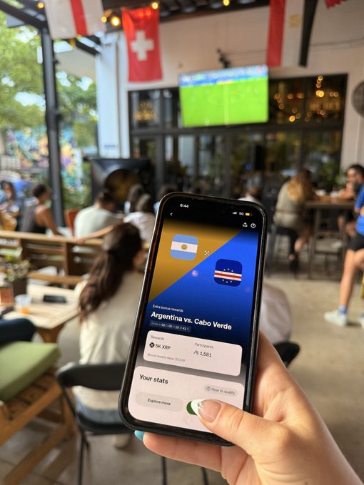 A person holds up a phone displaying a live betting slip for "Argentina vs. Cabo Verde" on the OKX app (SK XRP, potential return $1,581), while watching the match on a TV at a lively sports bar decorated with Swiss flags.