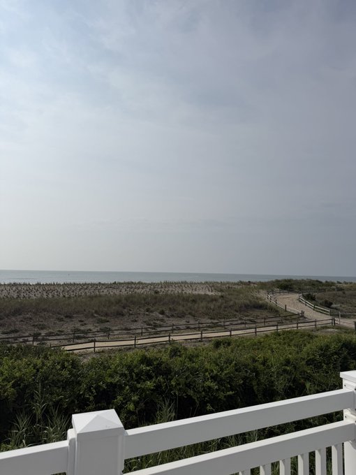 A serene coastal view with sandy dunes, greenery, and a calm sea beyond a white wooden railing—evoking the peaceful, carefree lifestyle the poster contrasts with their return to office ("cube") work. No visible text, numbers, or tickers in the image.