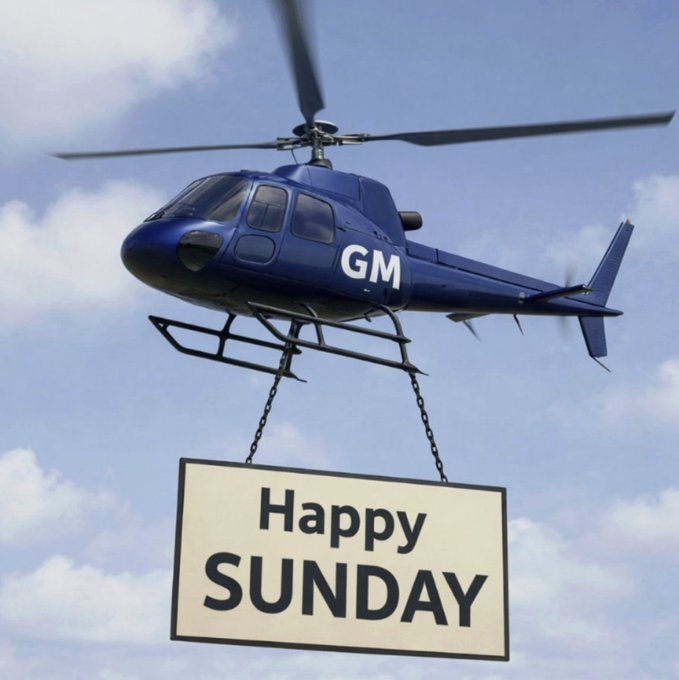 The image features a blue helicopter flying through a cloudy sky, with a banner attached to its side that says "Happy Sunday" written in black text. The banner also displays the words "GM Legends Fueling up for the next sprint". This suggests that the helicopter is being used as an advertisement or promotional tool for GM, possibly indicating their presence at an event or gathering on Sunday.