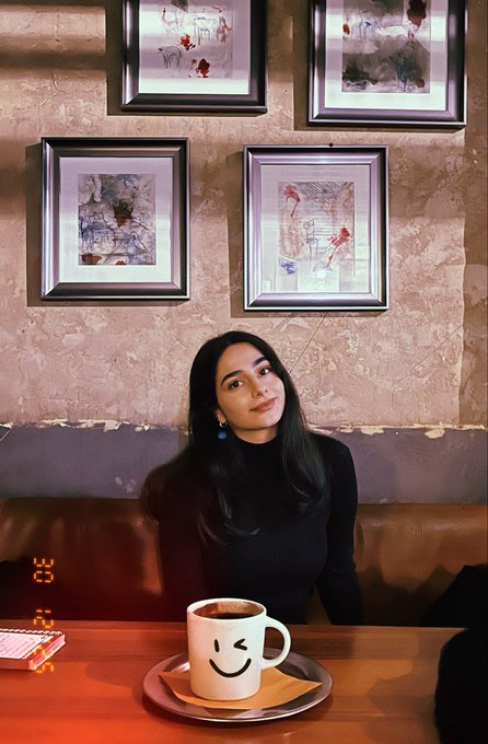 A young woman with long dark hair wearing a black turtleneck sits at a wooden table in a cafe with vintage framed artwork on the wall behind her; a white smiley-face coffee mug sits on the table in front of her.