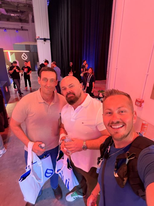 Three men pose for a selfie in a large event space with pink lighting; the man in the middle wears a white polo shirt, and they are holding white tote bags.