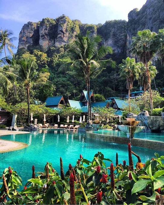 The image shows a beautiful tropical resort with a large swimming pool surrounded by palm trees and lush greenery. There are several buildings visible near the pool area, including a hotel that appears to be part of the resort. The scene conveys a sense of relaxation and leisure as guests can enjoy the natural surroundings while staying at the resort.