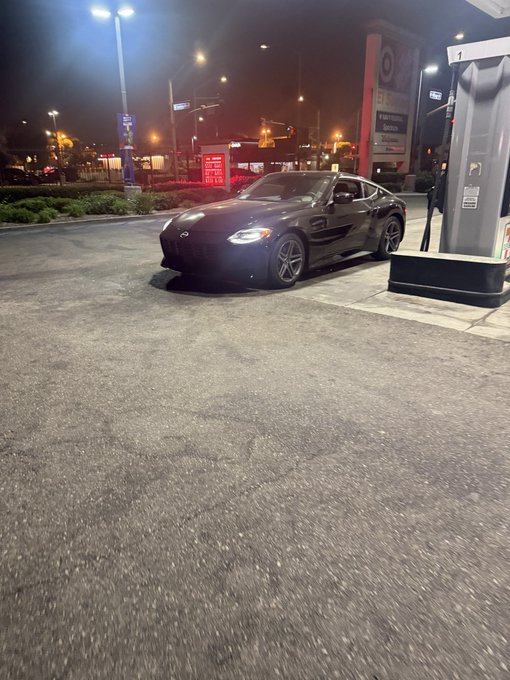 A black sports car is parked at an EV charging station at night, with bright overhead lights illuminating the scene.