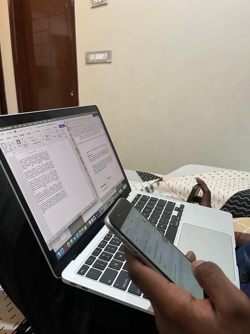 A person works on a laptop displaying a Microsoft Word document while holding a smartphone, possibly reading or messaging.