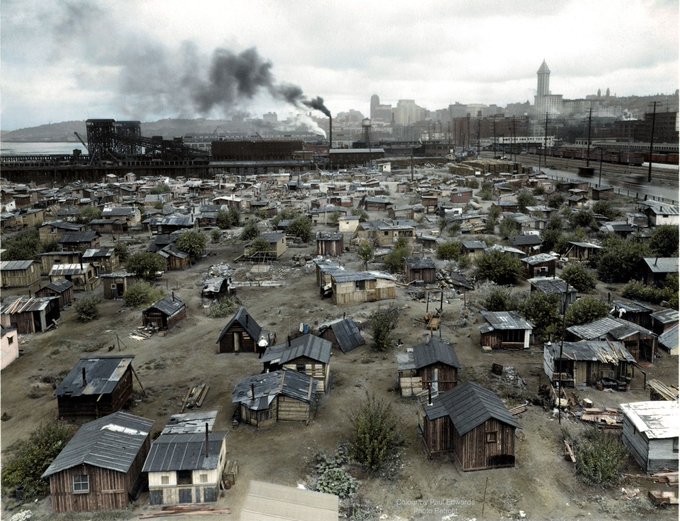 A vast shantytown ("Hooverville") of makeshift wooden shacks sprawls across the foreground beneath heavy industrial smokestacks billowing black smoke, illustrating the severe poverty and urban squalor that defined "Trumpvilles 1929" during the Great Depression.