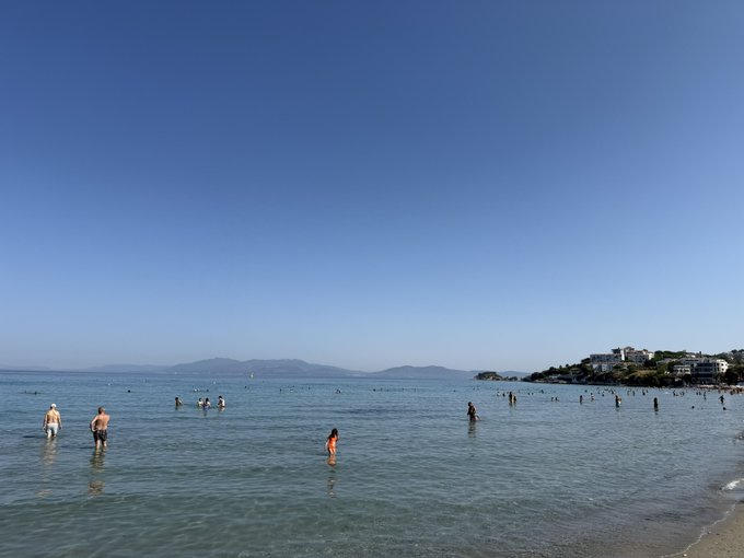 A serene beach scene shows numerous people wading in calm, clear ocean waters under a vast blue sky, with a distant shoreline and hillside buildings visible on the right. The image of swimmers enjoying a popular beach contrasts with the post's humor about struggling to get engagement ("GM" replies) on social media, suggesting the user feels they're missing out on attention while others receive abundant interactions.