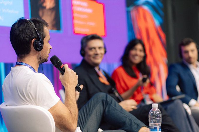 A panel discussion features four people wearing headsets, with one man in a white shirt speaking into a microphone while others listen, set against a purple backdrop.