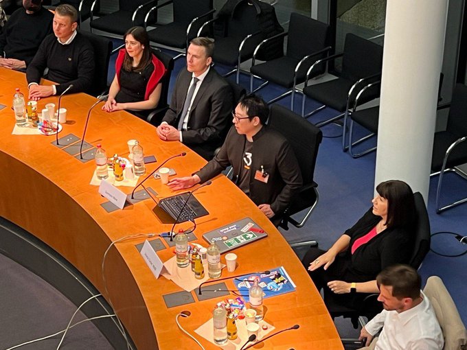 A panel discussion takes place at the German Bundestag, where Samuel Lee (Excellion) sits alongside German politician Joana Cotar and other officials, advocating against the sale of Bitcoin reserves. The image captures a formal parliamentary session with name placards, microphones, and refreshments arranged on the curved table where the speakers are seated.