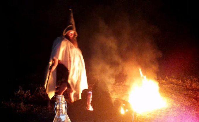 A man in a tall pointed hat and light robe stands barefoot near a large bonfire at night, with smoke billowing around him and a lantern visible in the foreground. The "live broadcast in progress" caption (直播正在进行中) combined with the contract address suggests this image is being used as a surreal, meme-like backdrop for a livestream tied to a meme token on the Solana blockchain.