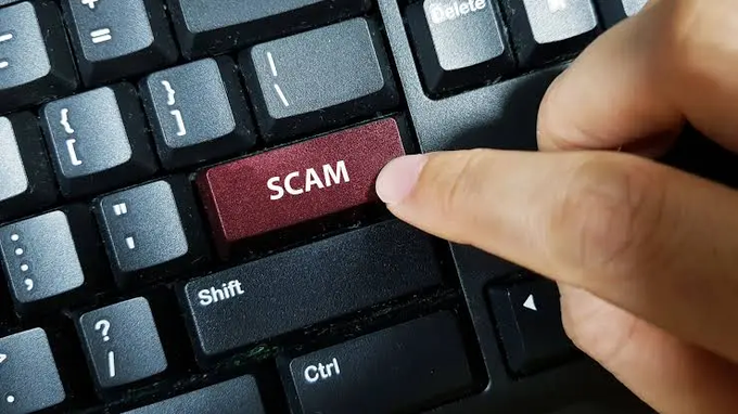 A close-up photo of a finger pressing a red 'SCAM' key on a black computer keyboard.